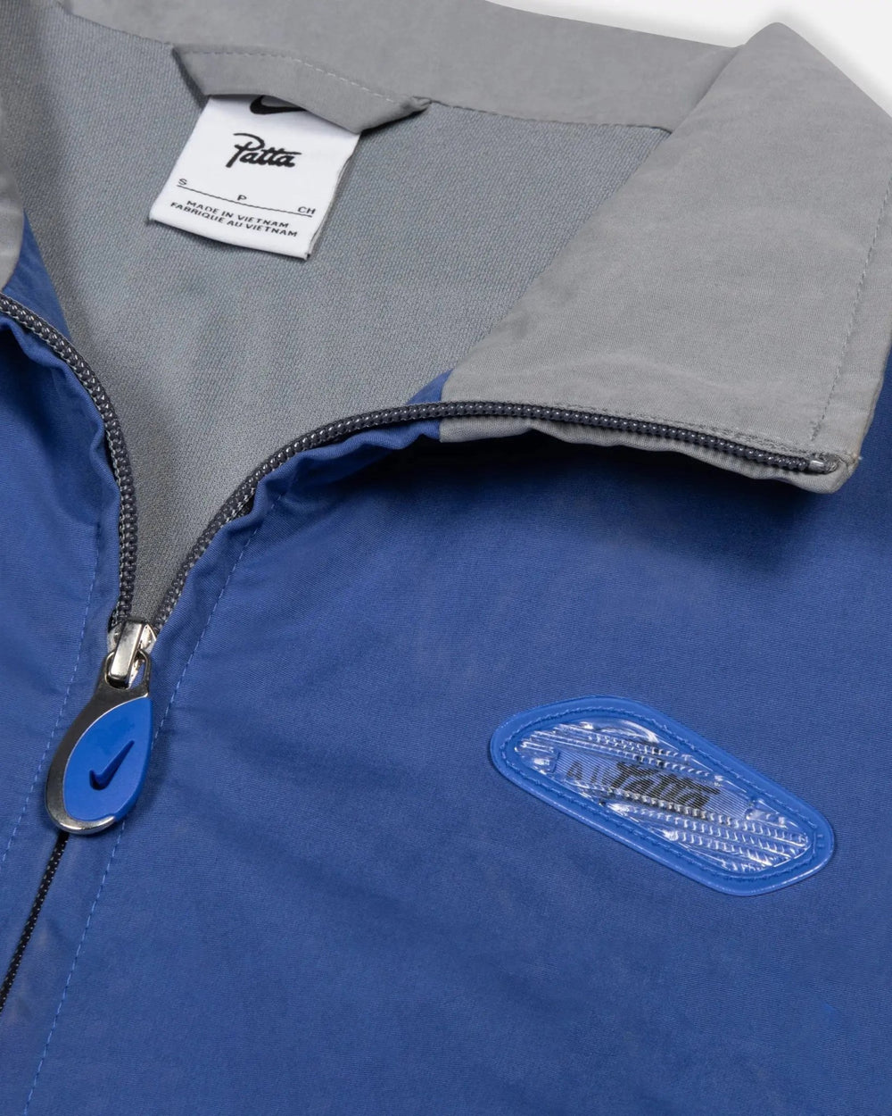 Collar and chest detail of the Patta x Nike NRG Track Jacket in sapphire blue with lenticular badge and zip puller.